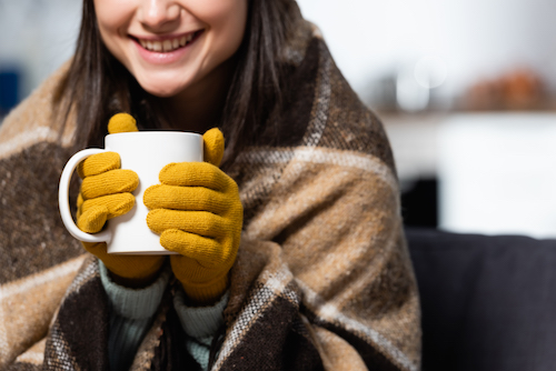 cropped view of woman wearing knitted gloves, wrapped in plaid blanket, holding cup of warm tea at home